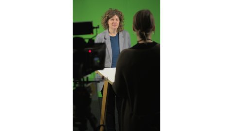 Ulrike Peter steht vor einem Greenscreen. Im Vordergrund eine Kamera und eine Person. Ulrike Peter steht vor einem Greenscreen. Im Vordergrund eine Kamera und eine Person.